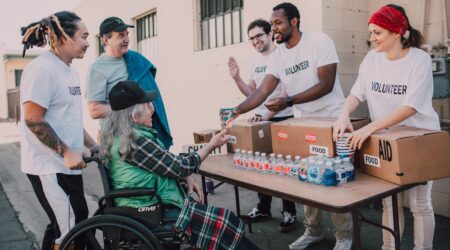 A group of volunteers serve a beverage to an older man in a wheelchair