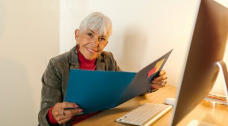 An older woman smiles while holding a file at a computer